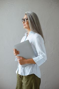 Confident mature Asian business lady wearing elegant blouse holds contemporary laptop standing near grey stone wall in studio side view