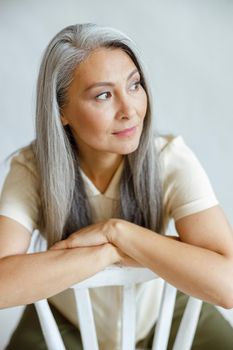 Pretty middle aged Asian lady with straight grey hair sits backwards on chair on light background in studio. Mature beauty lifestyle