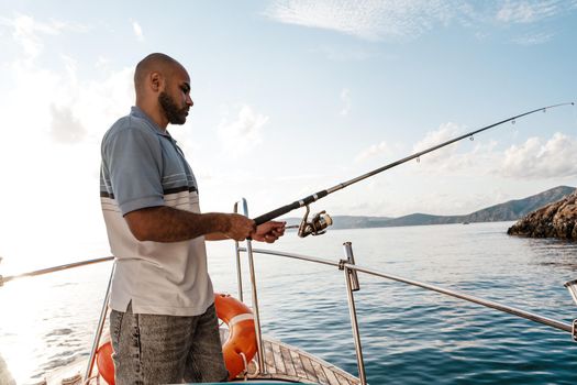 Young african american man standing with fishing rod on a sailboat fishing in open sea on sunset, close up