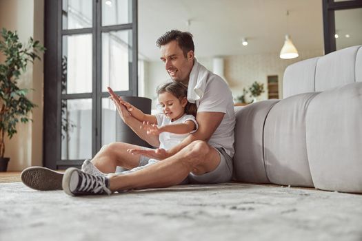 Low angle of smiling man with little girl sitting on floor in living room and having fun