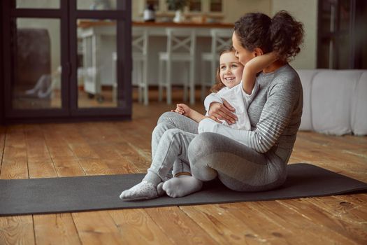 Cheerful woman is cuddling with girl while doing gymnastics on mat in cozy living room