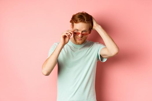 Handsome young redhead man in sunglasses, brushing hair with hand and looking smug and confident at camera, standing over pink background.