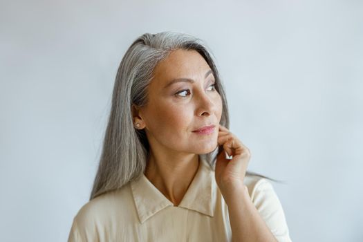 Thoughtful grey haired woman in stylish shirt looks aside on light background in studio. Mature beauty lifestyle