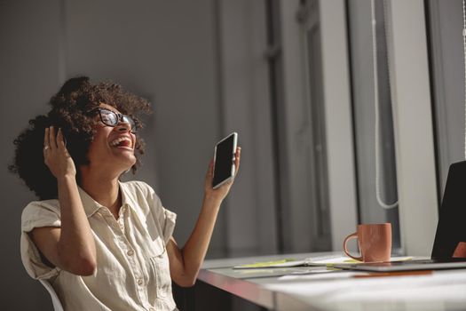 Happy Afro American female sitting in the coworking headphones and holding mobile phone