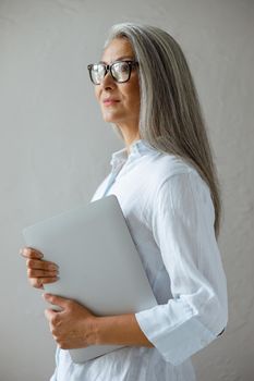 Cheerful middle aged Asian business lady wearing stylish shirt holds modern laptop standing near grey stone wall in studio side view