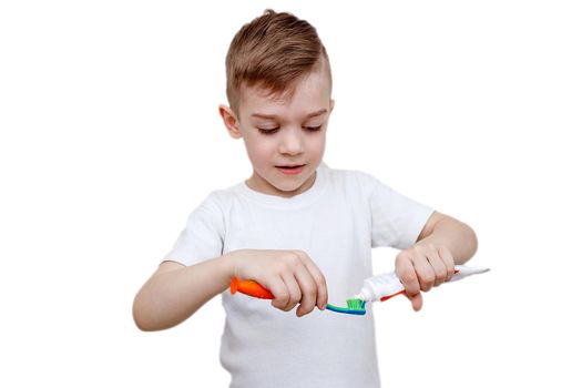 little five-year-old boy in white t-shirt squeezes toothpaste on brush and prepares to brush his teeth. Health care, hygiene and childhood concept. Caries prevention. Isolated on white background