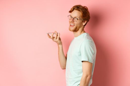 Handsome young man standing in profile and talking on speakerpone, record voice message, turn head at camera and smiling pleased, standing over pink background.