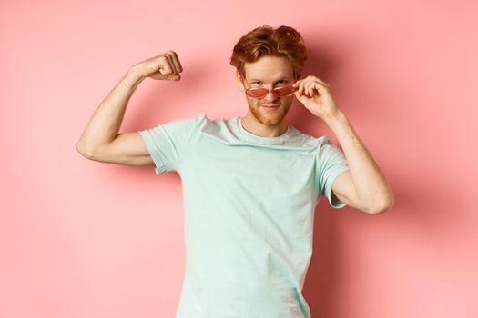 Confident young man with red hair, wearing summer sunglasses and t-shirt, showing strong and fit body muscles, flex biceps and staring cool at camera, pink background. Workout and gym concept