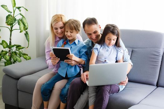 Happy family of four parents and cute little kids children enjoy using the tablet, watching cartoons, make internet video call or shopping online looking at computer screen sit together at home