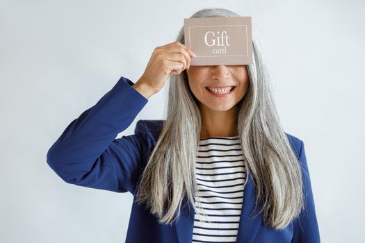 Smiling mature lady with loose silver hair in blue jacket holds gift card posing on light grey background in studio