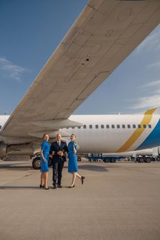 Full length shot of pilot standing together with two stewardesses in bright blue uniform in front of an airplane on a sunny day. Aircraft, aircrew, occupation concept