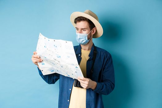Covid-19, pandemic and travel concept. Handsome young guy in face mask and summer hat looking at tourist map, travelling on vacation, standing on blue background.