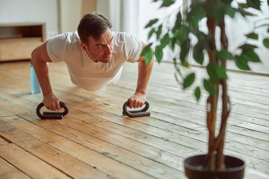 Athletic man is doing push ups while using special handles during workout in living room