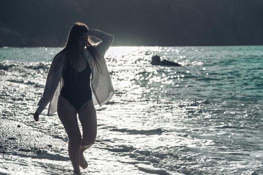 Selective focus. Happy carefree sensual woman with long hair in black swimwear posing at sunset beach. Silhouette of young beautiful playful positive woman outdoor. Summer vacation and trip concept.