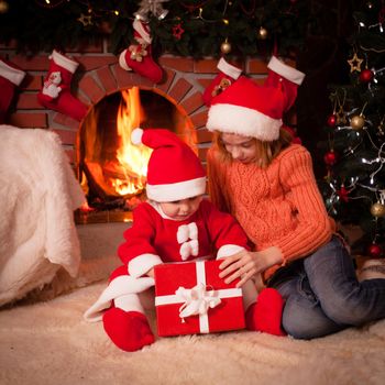 Children are sitting near fireplace and christmas tree with gift boxes.