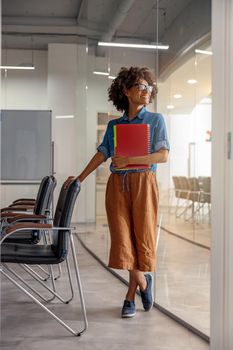 Full-length view of happy Afro American woman in glasses holding folders while waiting in the office, looking away