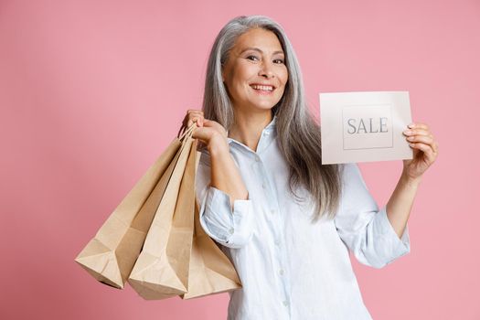 Beautiful middle aged Asian lady holds paper shopping bags showing card with word Sale on pink background in studio