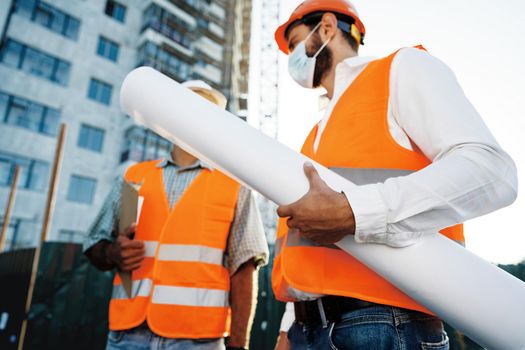 Two men in workwear and medical masks working with blueprints on object, close up