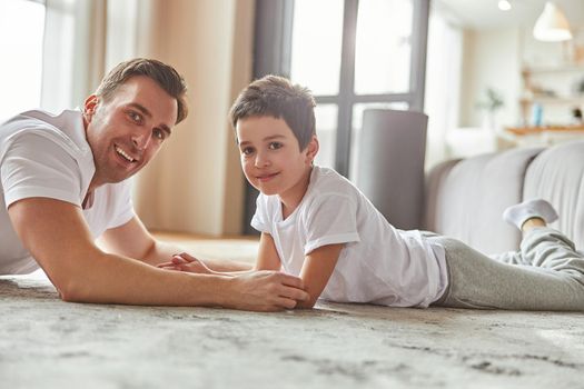 Waist up portrait of cheerful man lying on floor with cute boy while they are spending day together indoors