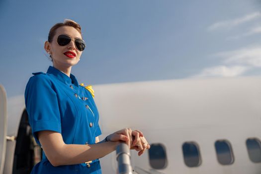 Portrait of pretty air stewardess in blue uniform and sunglasses looking away, standing on airstair on a daytime. Aircrew, occupation concept