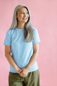 Happy silver haired middle aged Asian woman in blue t-shirt stands on pink background in studio. Mature beauty lifestyle