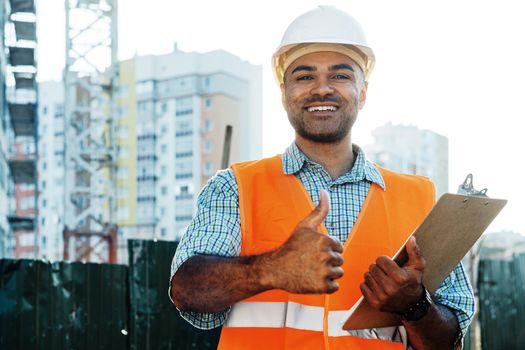 Young man engineer in workwear standing in construction site with clipboard, close up portrait