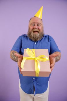 Emotional middle aged fat man wearing blue shirt and yellow hat stands on purple background in studio, focus on hands with gift box