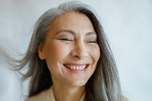 Joyful middle aged Asian female model with flying silver hair stands on light grey background in studio closeup. Mature beauty lifestyle