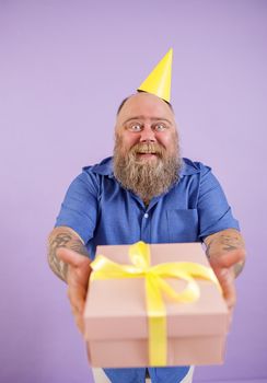 Happy bearded fat man wearing blue shirt and party hat presents gift box standing on purple background in studio