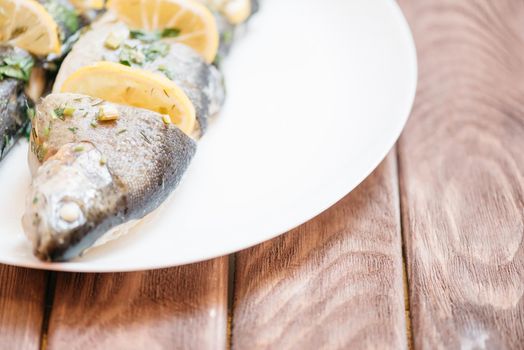 Baked fish trout dish with lemons and greenery on a white plate on wooden table.