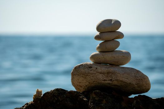 Balanced Pebbles Pyramid on the Beach on Sunny Day and Clear Sky at Sunset. Blue Sea on Background Selective focus, zen stones on sea beach, meditation, spa, harmony, calm, balance concept.