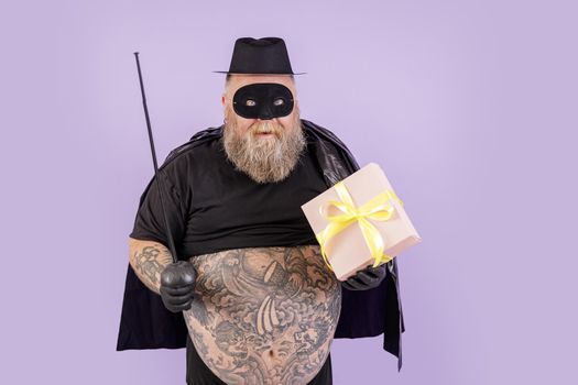 Happy middle aged gentleman with overweight wearing Zorro suit holds toy epee and gift box on purple background in studio