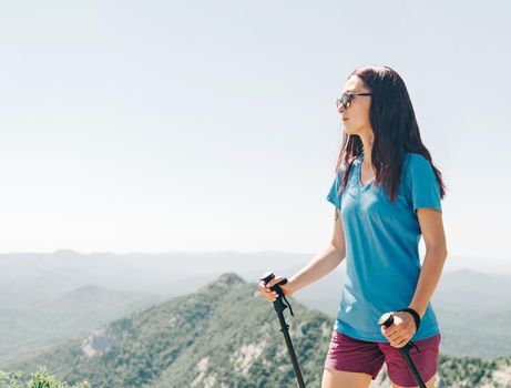 Active sporty young woman in a t-shirt and shorts walking with trekking poles in summer mountains outdoor.
