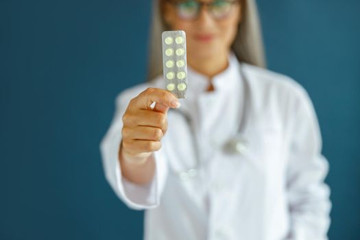 Middle aged lady therapist in white coat stands on blue background in studio closeup, focus on hand with blister of pills. Professional medical staff