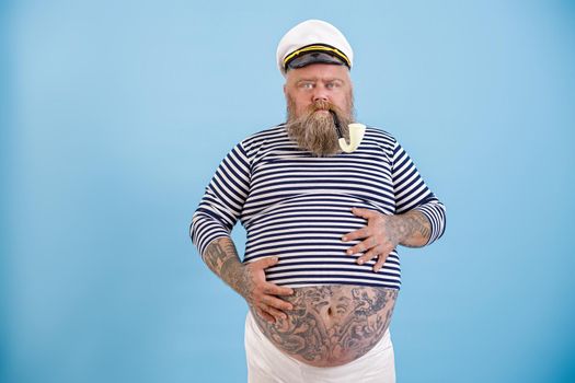 Middle aged obese man in sailor costume with smoking pipe holds hands on large tummy with tattoos on light blue background in studio