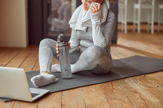 Cropped head of cheerful female sitting on mat and drinking water during workout on notebook