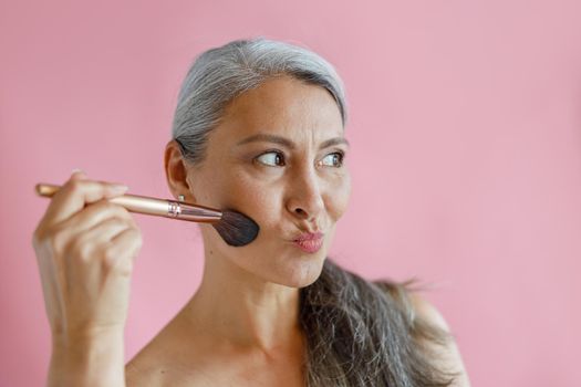 Funny hoary haired Asian lady applies makeup with cosmetic brush grimacing on pink background in studio. Mature beauty lifestyle