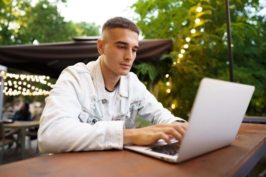 Young man sitting at table and typing on laptop keyboard while working in outdoor cafe, close up