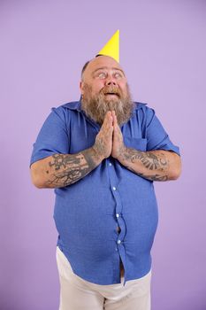 Funny mature bearded obese male person in blue shirt and party hat holds hands in praying gesture on purple background in studio