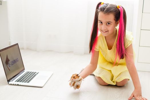 Cheerful young little girl with a pet hamster using laptop computer studying through online e-learning system at home. Distance or remote learning