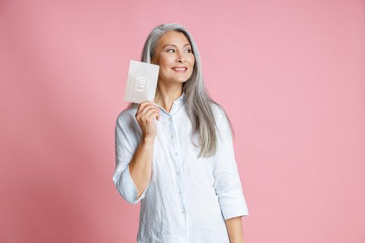 Pretty cheerful silver haired Asian woman holds gift card having ideas on pink background in studio. Shopping certificate