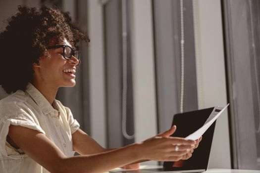 Smiling Afro American lady working with laptop while holding document and sitting near the window