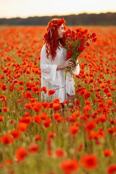 Portrait of beautiful redhead woman in wreath on green field with bouquet of poppies in summer sunset