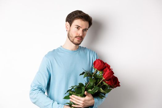 Handsome young man holding beautiful red roses for his lover on Valentines day, looking pensive, standing over white background.