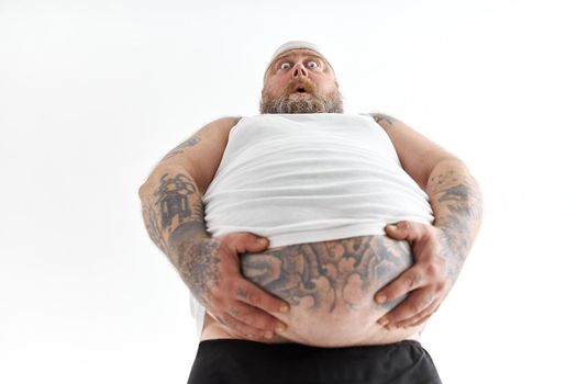 fat man with big belly and tattoes in sports wear is holding his stomach with shocked emotion