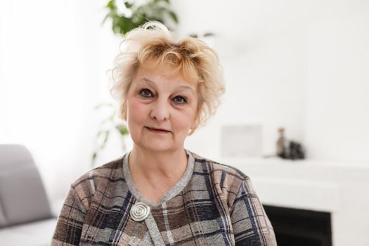 Attractive elderly woman sitting on couch at home, looking at camera, makes video call, communication distantly, modern wireless technologies concept