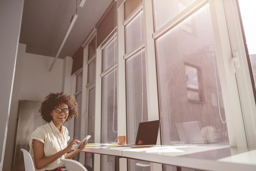 Happy pretty woman sitting at work desk and working with laptop while holding mobile phone