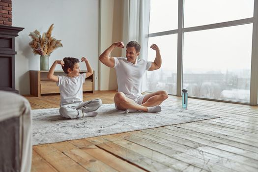 Happy man is sitting with boy on mat and showing biceps after doing workout together