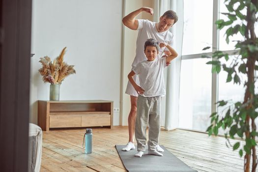 Full length portrait of cheerful father and son training on mat and demonstrating their muscles