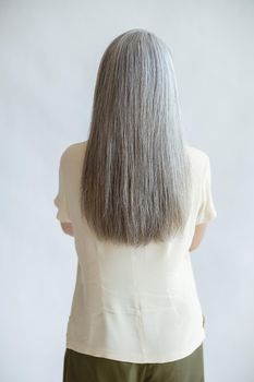 Lady in yellow blouse with long loose silver hair stands on light grey background in studio backside view. Mature beauty lifestyle